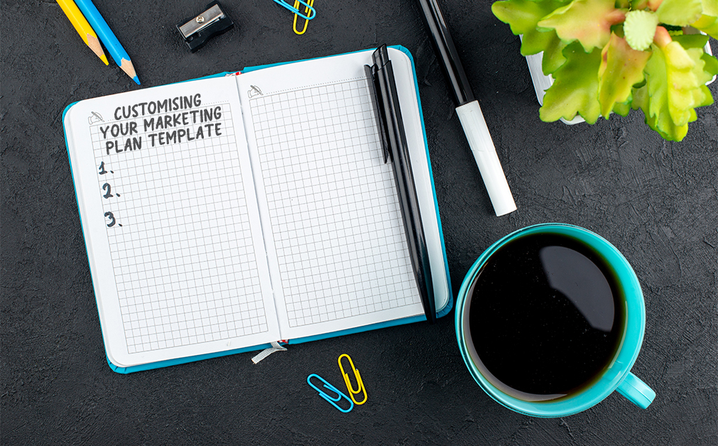 Overhead view of an open notebook on a dark desk. The left page has the heading “Customising Your Marketing Plan Template” with three numbered bullet points. Pens, paper clips, a turquoise coffee mug, and a small green plant are arranged around the notebook.