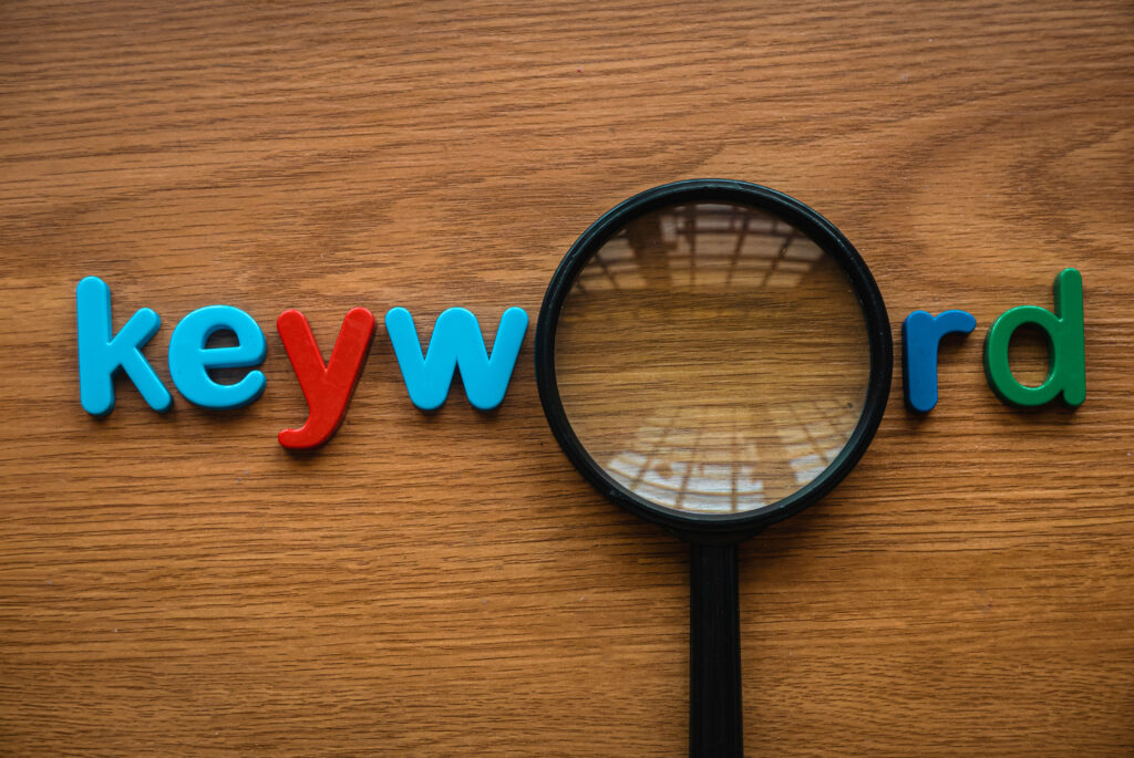 The image shows colorful plastic alphabet letters spelling the word "keyword," with a magnifying glass placed over the middle of the word. The background is a wooden surface, and the magnifying glass reflects a grid pattern.