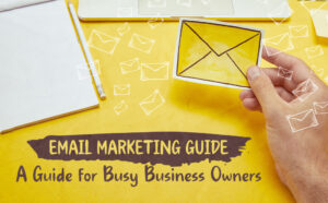 A hand holding a yellow envelope illustration over a yellow desk with a notepad, pencil, and laptop. White envelope icons float across the image. Text reads: "Email Marketing Guide – A Guide for Busy Business Owners."