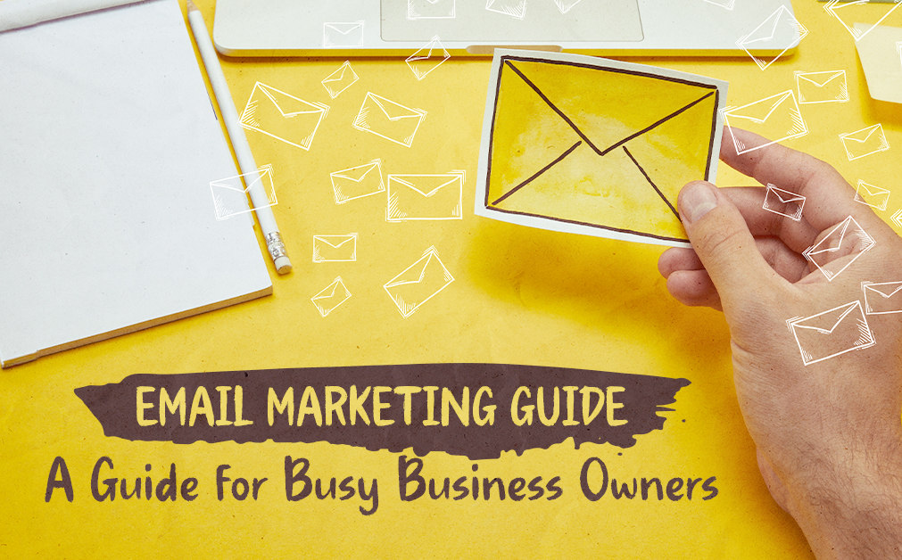 A hand holding a yellow envelope illustration over a yellow desk with a notepad, pencil, and laptop. White envelope icons float across the image. Text reads: "Email Marketing Guide – A Guide for Busy Business Owners."